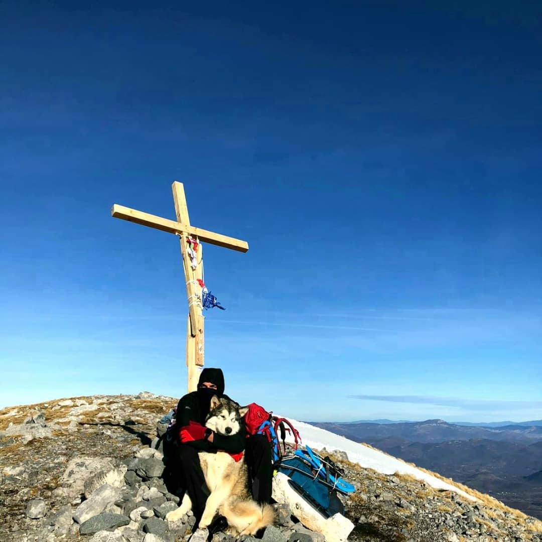 a man named grga brkic and his 8 month old alaskan malamute, north, sitting on a mountain top in croatia in front of a wooden cross