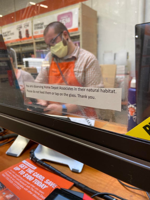 Home Depot workers behind plexiglass with a sign taped on that says "you are observing Home Depot Associates in their natural habitat. Please do not feed them or tap on the glass. Thank you."