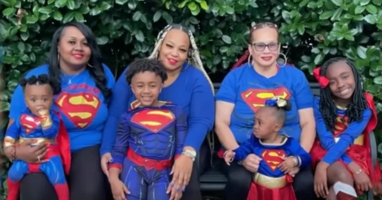 three women and four children, including 6-year-old kayden reid and 2-year-old kaylee reid, sitting on bench outside as they smile and pose in superman shirts or costumes