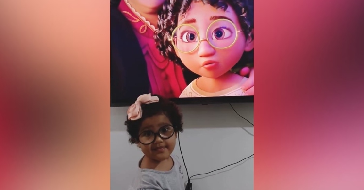 a 2 year old girl named manu araújo marques smiling as she stands next to a tv showing mirabel from the disney film "encanto"