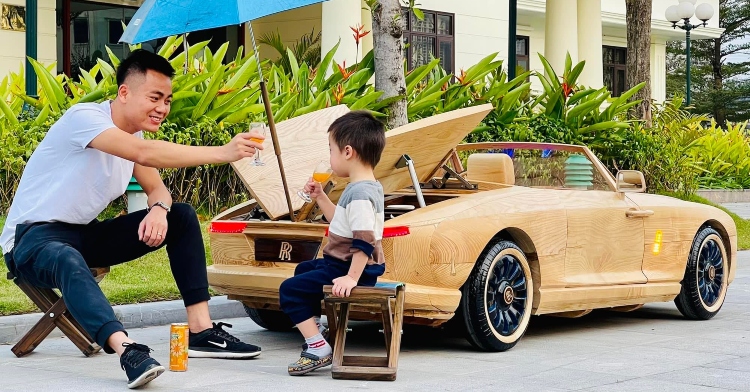 a man named trương văn dạo toasting a glass with an orange drink with his little boy as they sit outside next to a mini wooden replica of the rolls-royce boat tail that trương made