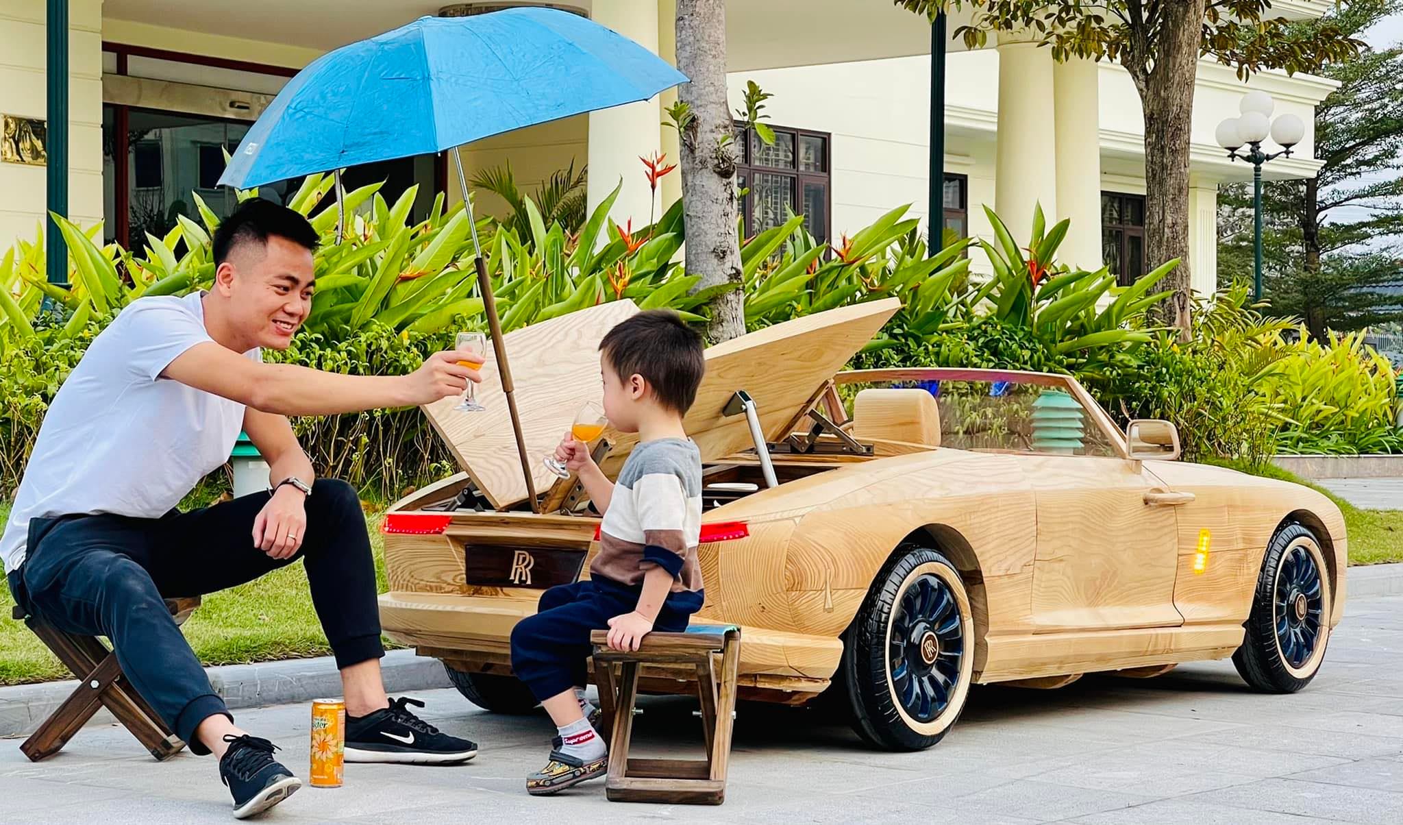 a man named trÆ°Æ¡ng vÄƒn dáº¡o toasting a glass with an orange drink with his little boy as they sit outside next to a mini  wooden replica of the rolls-royce boat tail that trÆ°Æ¡ng made