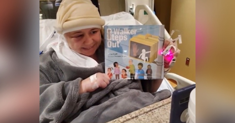 a woman named kd meucci who had brain surgery the day before sitting in a hospital bed as she smiles and holds up the book "mr. walker steps out" by lisa graff