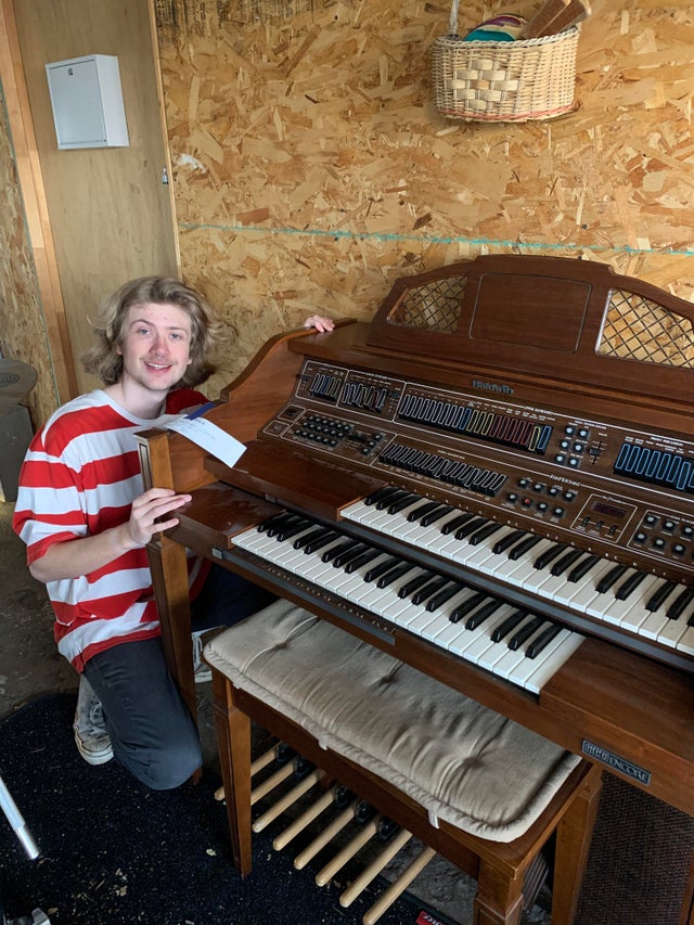 a man squatting as he smiles and poses with a baldwin organ he purchased at a thrift store 