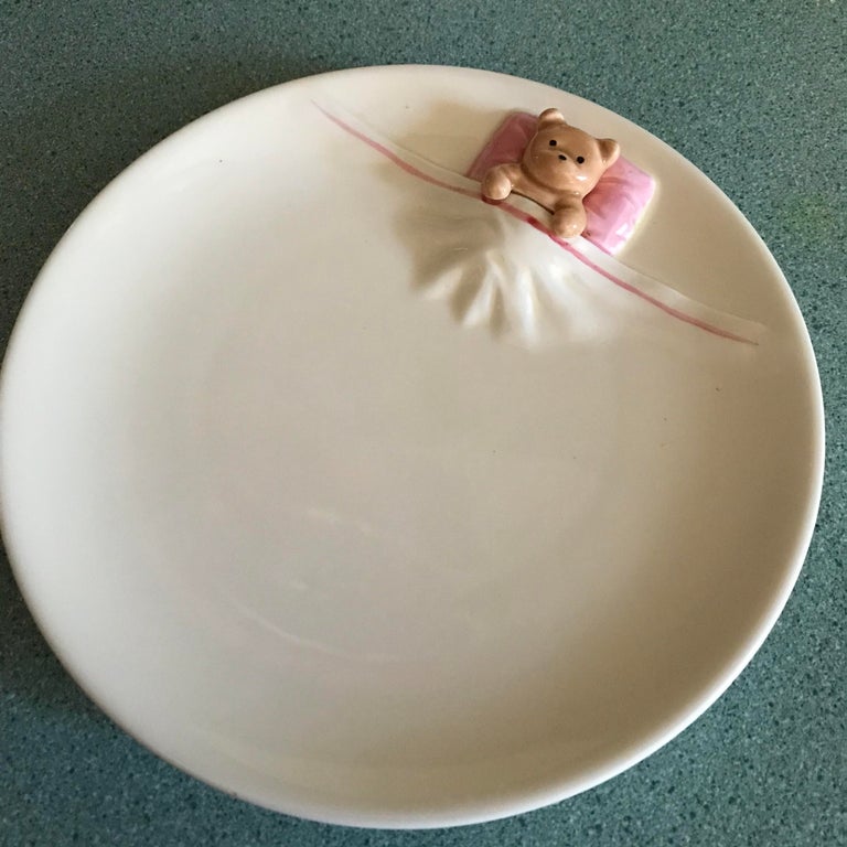 a cream colored plate made to look like a sheet that the small decorative bear is using as it lays its head on a pillow 