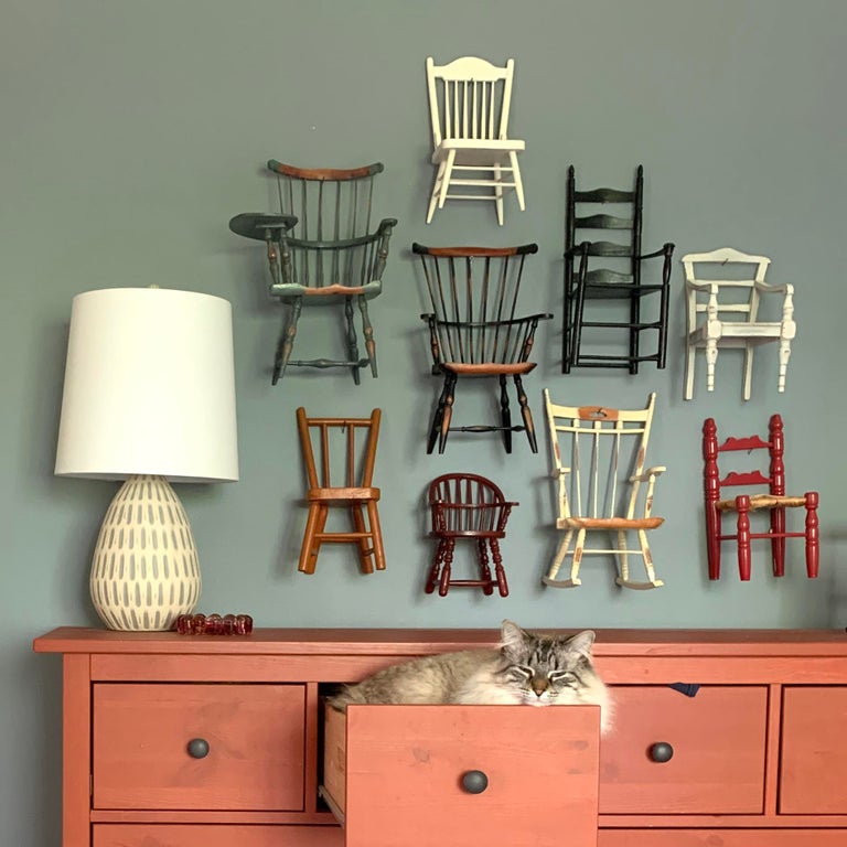 a cat sleeping in an open dresser drawer with nine miniature chairs hung on the wall above it