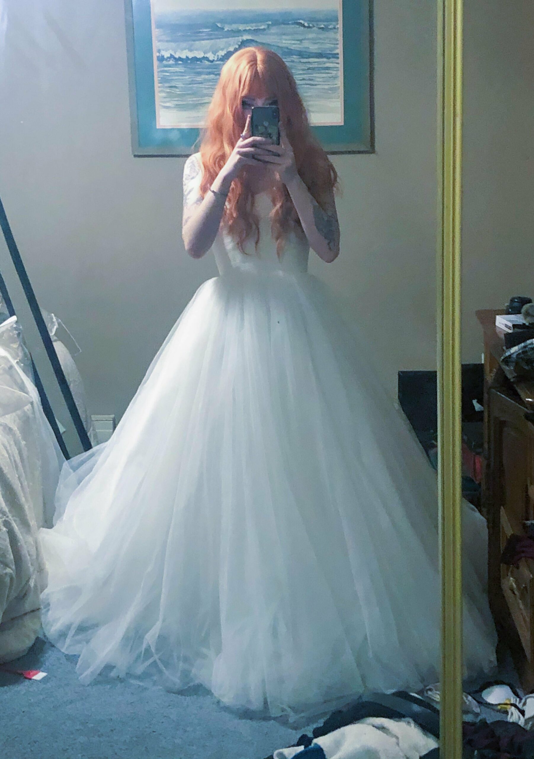 a woman with light orange hair taking a picture of herself in a mirror while wearing a wedding dress she purchased from goodwill