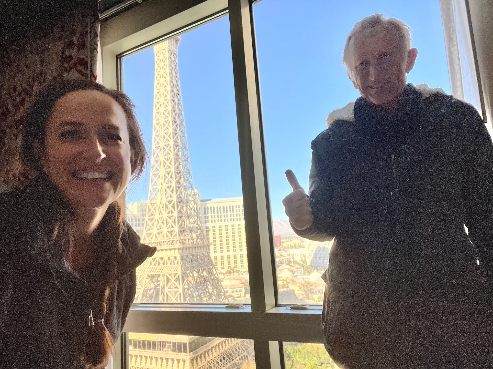 a woman named vanessa mitchell-delomotte and her dad, patrick mitchell, posing for a selfie with the view of the las vegas eiffel tower in the background as seen through a window 