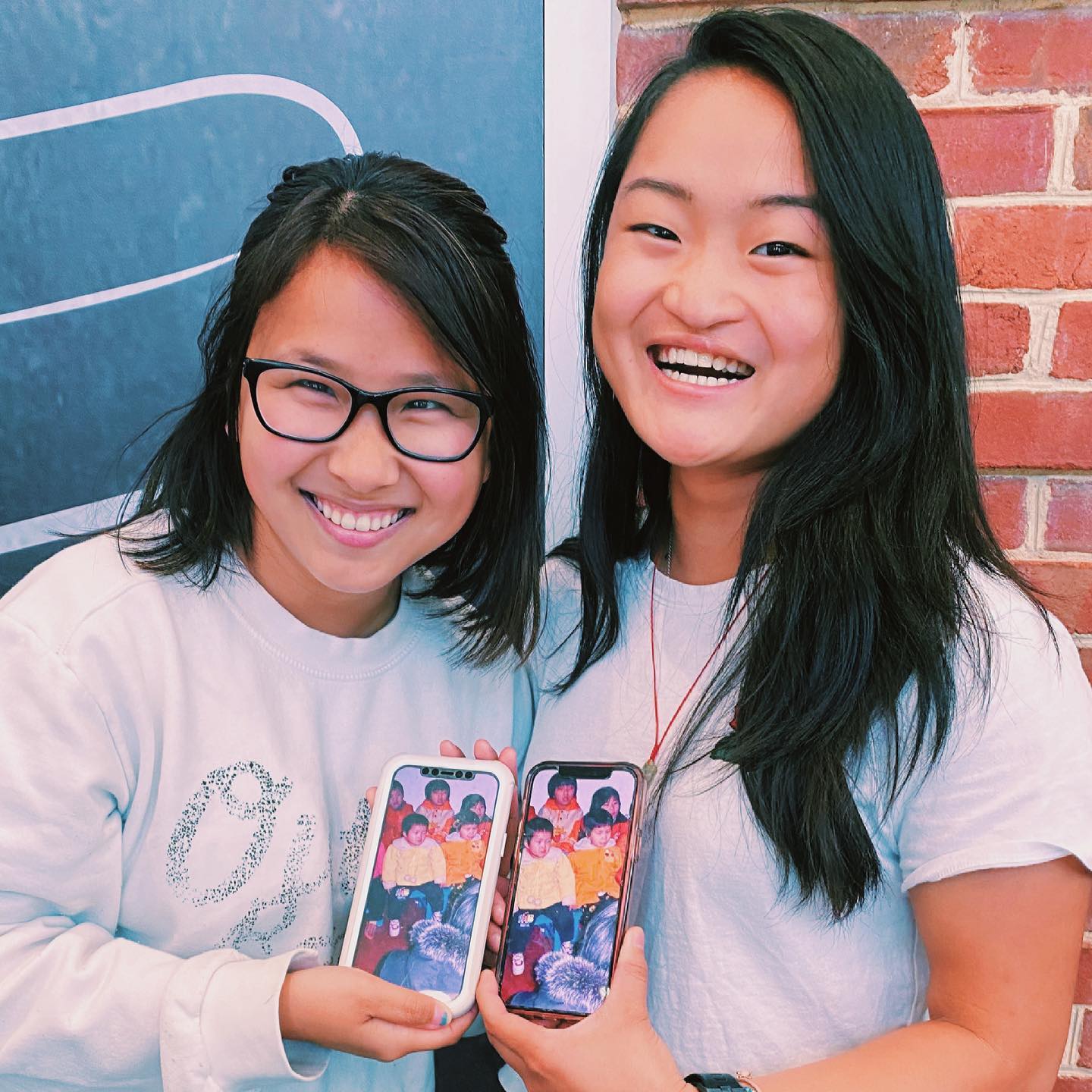 two young women named ally and ruby smiling and posing with identical photos of themselves in a chinese orphanage when they were kids on their phones