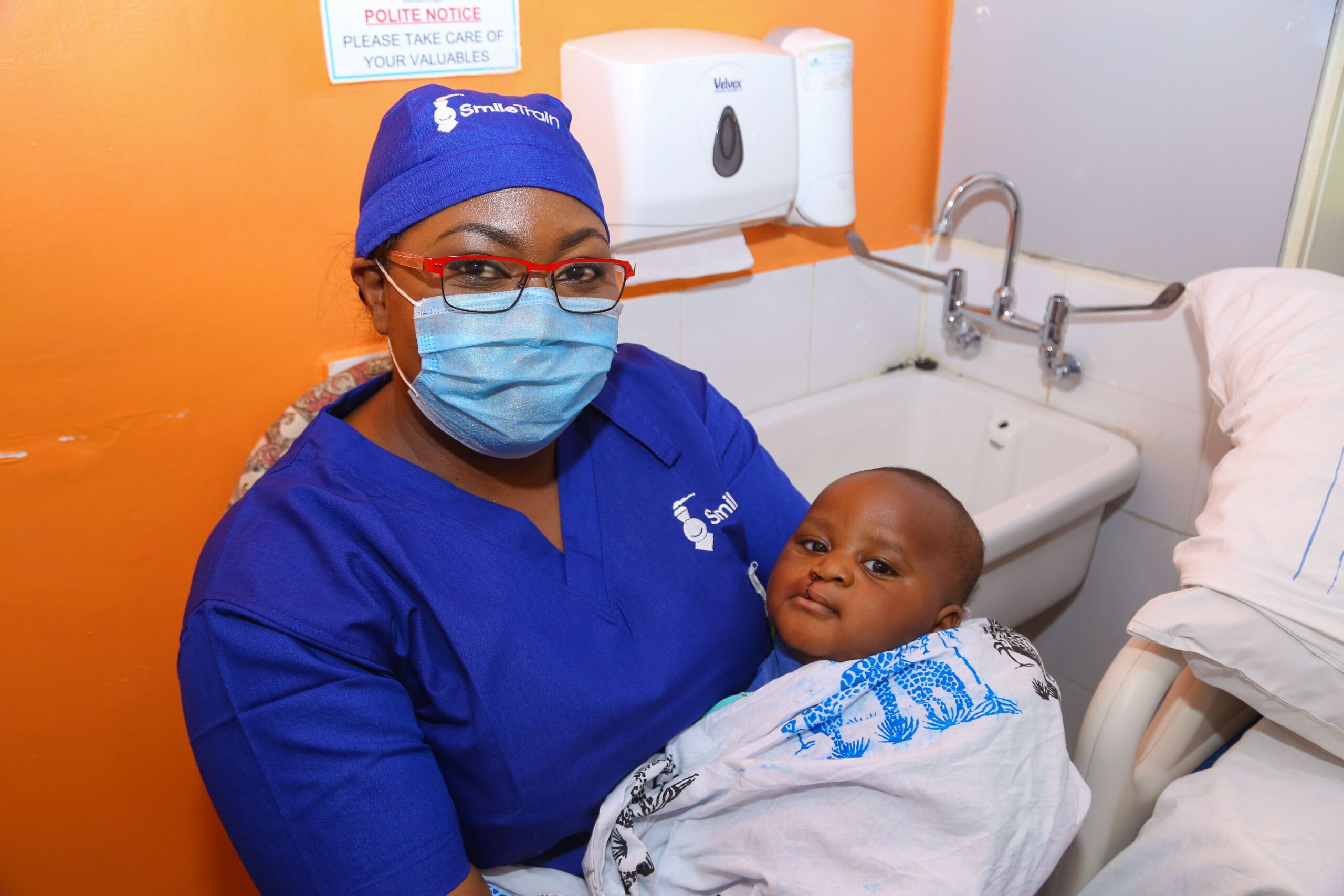 a surgeon named dr. amanda mulango wearing a face mask as she sits and holds a baby with a cleft