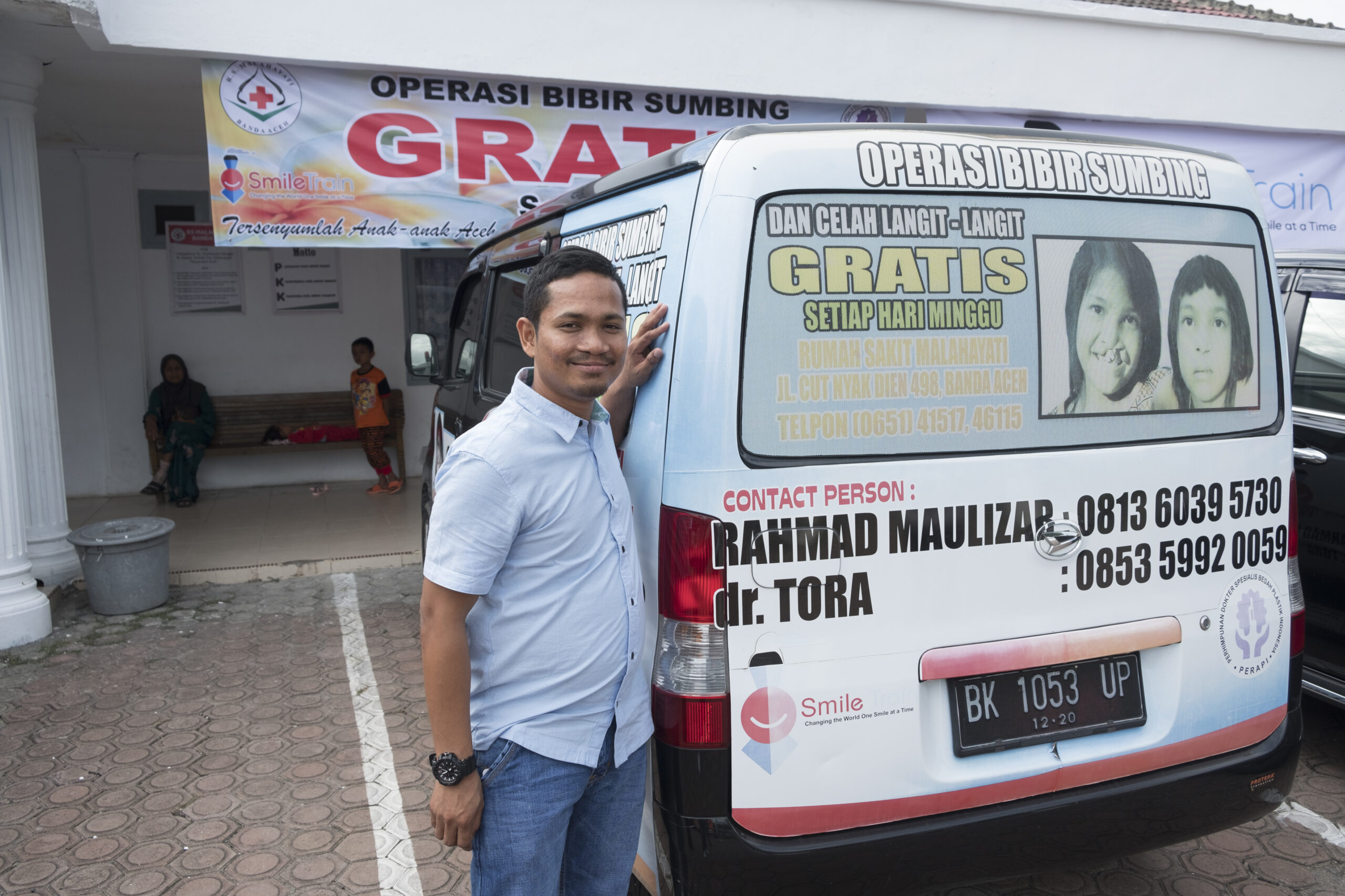 a man named rahmad smiling as he poses next to a van with before and after photos of children who received cleft surgery through the help of smile train 