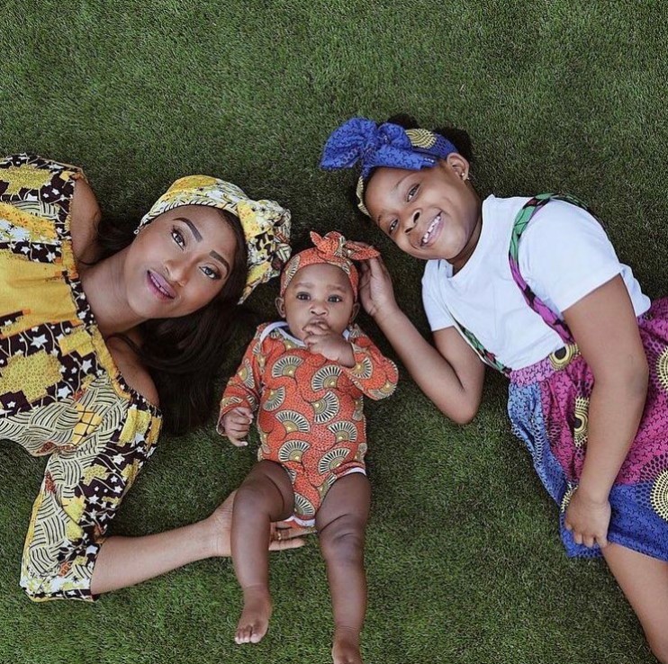 a woman, her baby girl, and her other young daughter looking up as they pose while laying on grass while the baby wears clothes from ade + ayo
