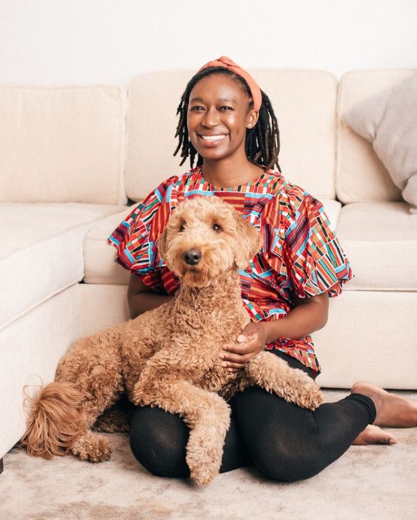 a woman named temidayo adedokun smiling as she sits on the floor in front of a couch with a large, curly haired dog on her lap