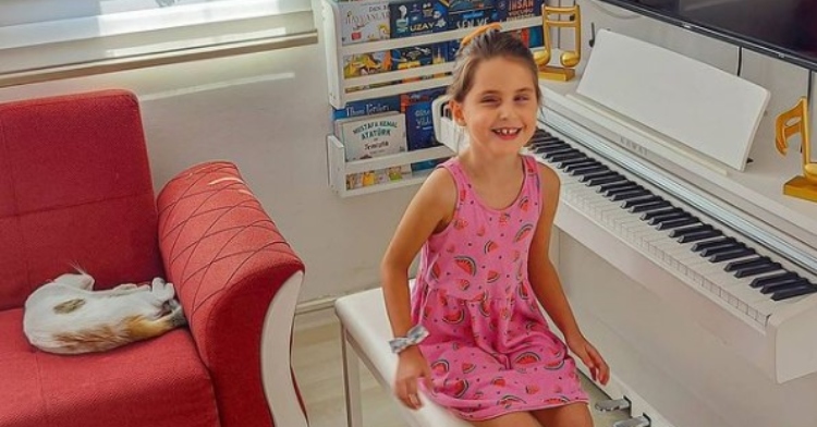 7 year old little girl named ipek nisa göker smiling as she sits on a bench next to a white piano and a sofa with a cat sleeping on it