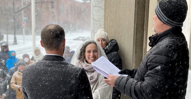 a bride named sally faulkner and a groom named adam irujo standing outside for their wedding ceremony as a crowd of their family and friends stand nearby and watch