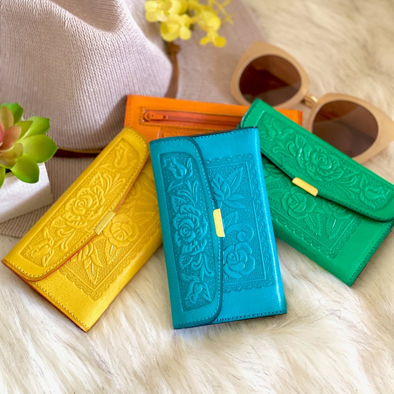 an assortment of handmade leather wallers in orange, yellow, blue, and green sat on a furry rug next to a hat and sunglasses by etsy user salylimonusa