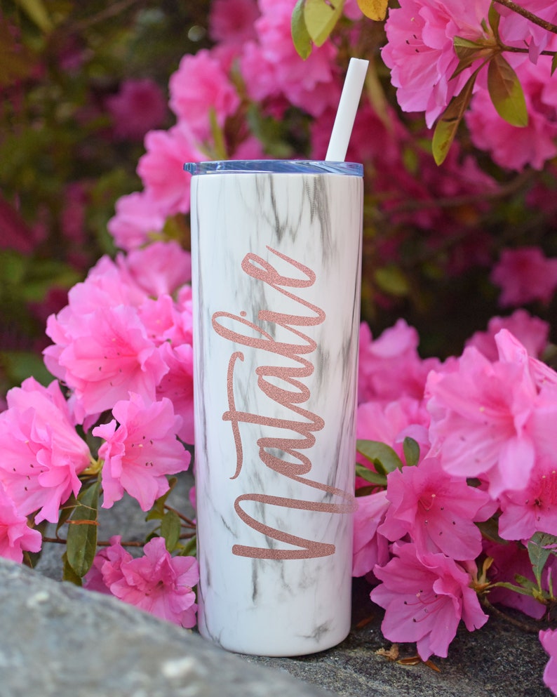 a black and white marble tumbler made by etsy user thewhiteinvitegifts that has the name "natalie" on the side and is placed outside in front of pink flowers