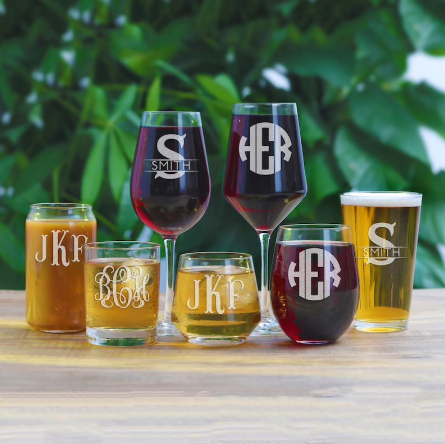 various types of drinking glasses with wine or beer that each have a persoanlized monogram, each of which are sat outside on a table and are made by etsy user yaypersonalizedgifts