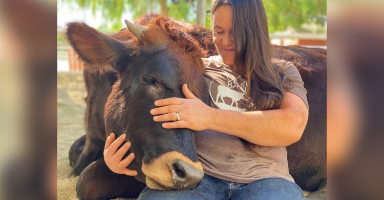 a woman named ellie laks smiling as she sits and hugs a sitting cow on her farm called the gentle barn