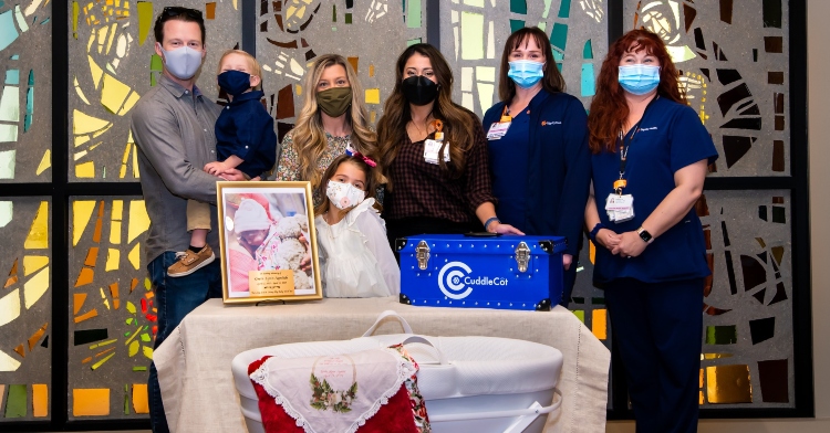 a family of three posing with four hospital staff members next to a photo of their recently passed new born, a bassinet, and the cuddle cot that the infant was placed inside of