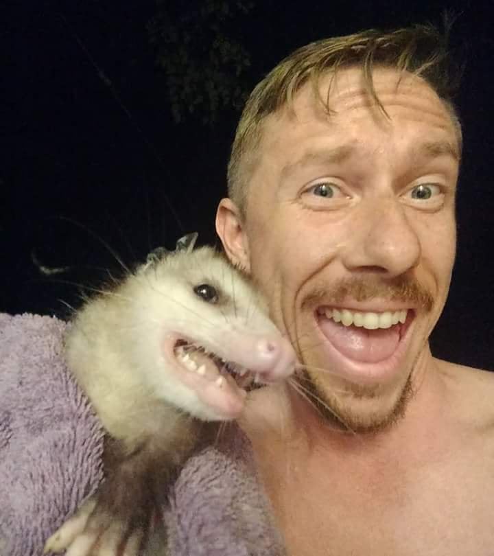 closeup of a shirtless man smiling for a selfie with the opossum he's carrying in a towel to take back outside after it got inside his house
