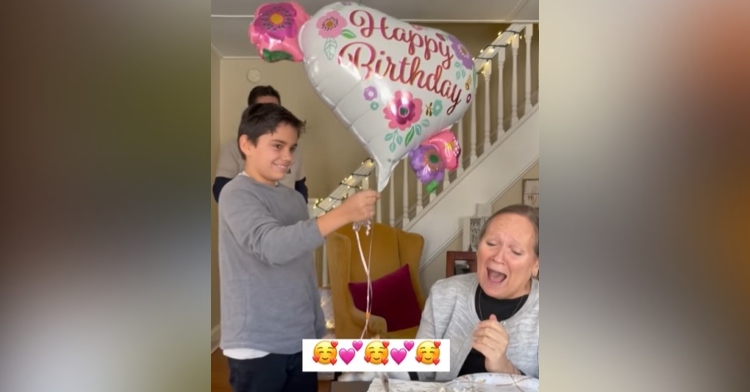 a 70 year old woman with her mouth agape from excitement as her grandson hands her a heart shaped balloon that says “happy birthday” on it