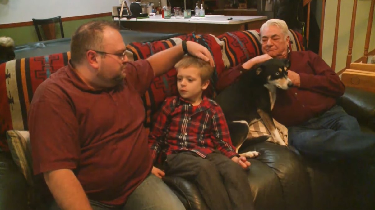 a man named brad harbert, his son, their dog named roxy, and brad's dad all sitting together on a couch in their living room
