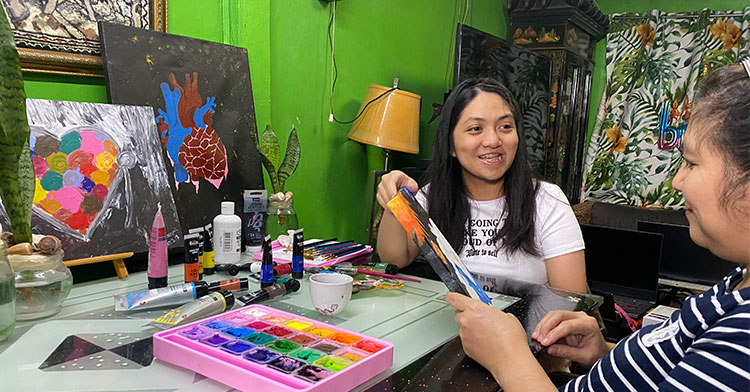 artist surrounded by art supplies and a few paintings at table with her mom