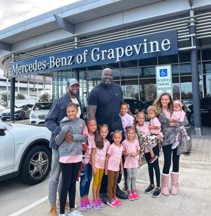 the collins family, which includes a mom, dad, and nine kids, all smiling and posing with shaquille o'neal outside of the mercedes-benz of grapevine