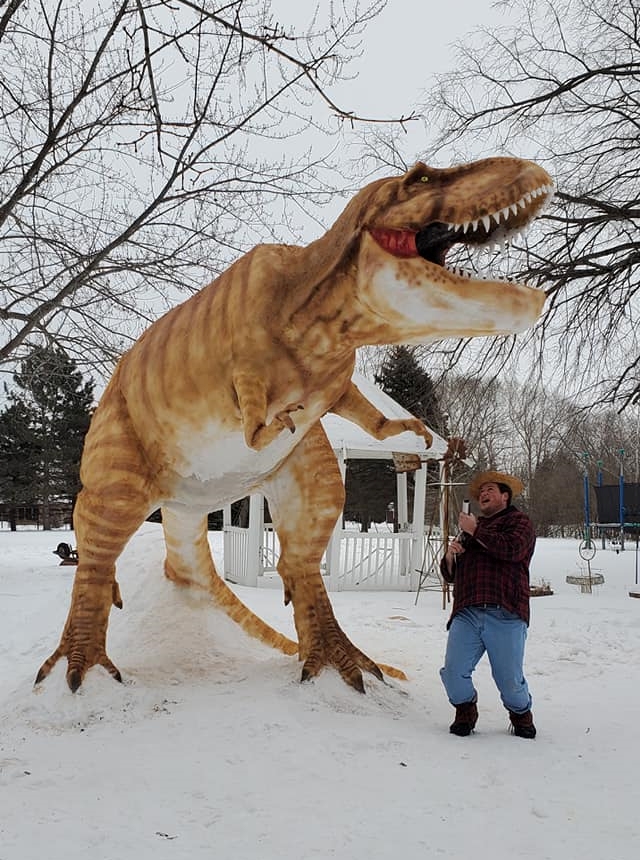 a man named paul larcom playfully looking up as he points a gun at his 12 foot tall t-rex snow sculpture