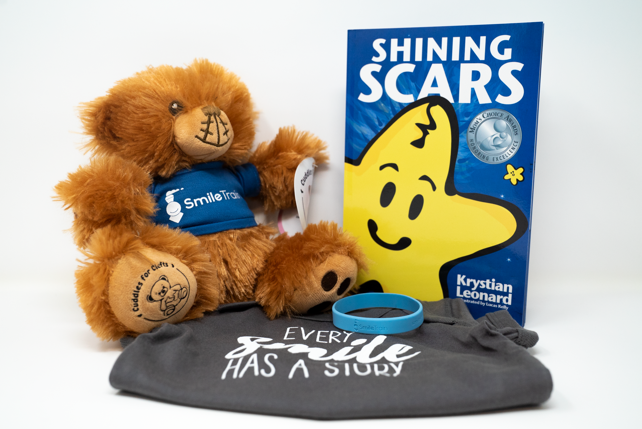 a care package from cuddles for clefts in partnership with smile train called smile train cuddle pack that features a stuffed animal, a book called "shining scars," a onesie that says "every smile has a story," and a blue bracelet