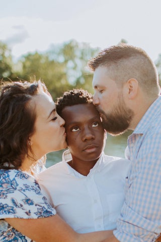 closeup of a woman and a man each kissing the side of their adopted thirteen year old son's face as he rolls his eyes