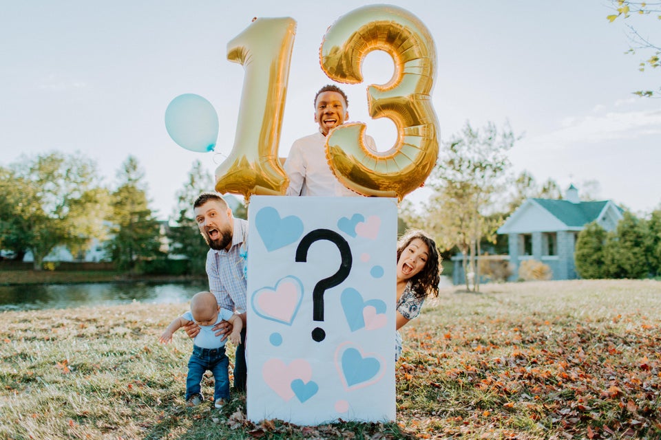 a man and a woman peaking from behind a box with a question mark with a baby as a teenage boy smiles while standing inside the box along with a "1" and a "3" balloon 