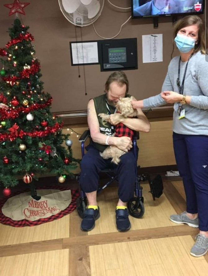 a nurse standing by as a man cries and holds his dog while sitting next to a christmas tree 