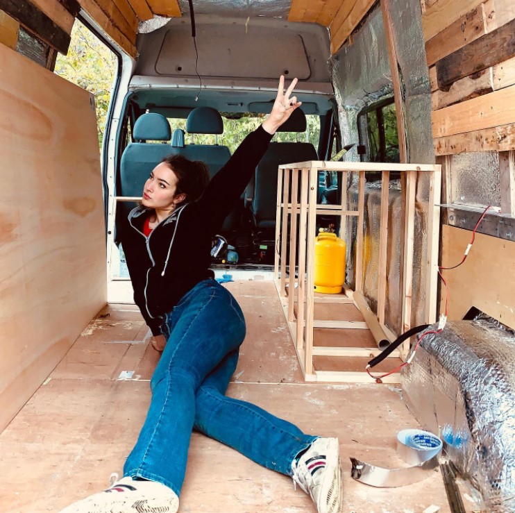 a woman named ame posing inside of her van that she's renovating by sitting down with her legs stretched out as she gives a peace sign