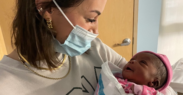 a woman named stela niznik wearing a face mask and smiling as she holds and looks down at the baby girl she delivered named darlie stela