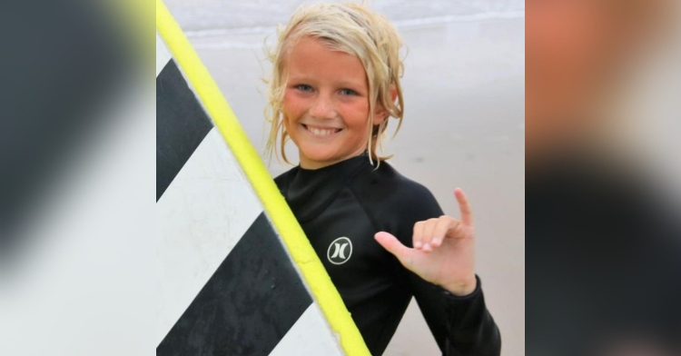 closeup of an 11-year-old boy named carter doorley smiling as he holds his striped surfboard and give the “hang loose” sign with his hand