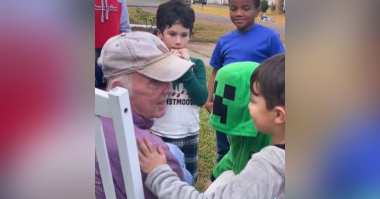 91-year-old man named gene mcghee sitting in a white chair in his front lawn as the students of a daycare across the street stand around and talk to him, one of which is a little boy who has one of his hands placed on gene’s shoulder as he speaks to him