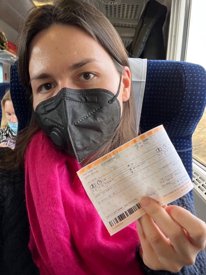 a ukrainian woman who is wearing a black face mask sitting on a train in poland as she holds up her ticket to show it didn't cost her anything