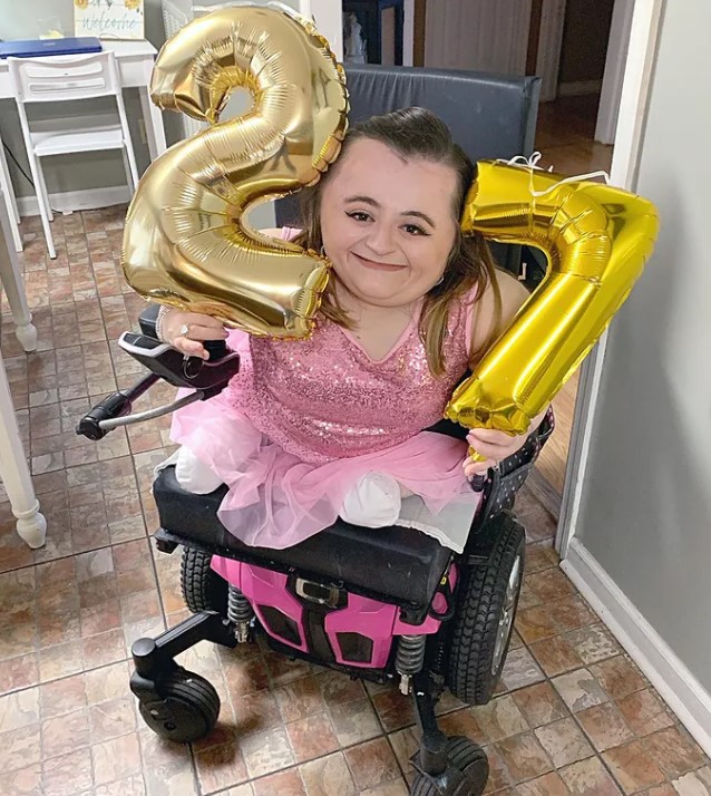 27-year-old hannah trye smiling as she sits in her wheelchair and holds two balloons, one that's the number "2" and the other that's the number "7"