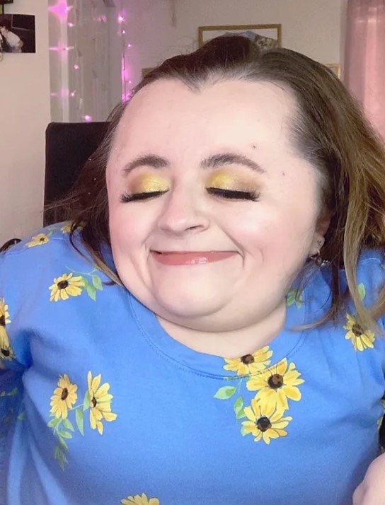 closeup of hannah trye who is wearing a blue shirt with yellow flowers and is smiling with her eyes closed to show off her golden eyeshadow 