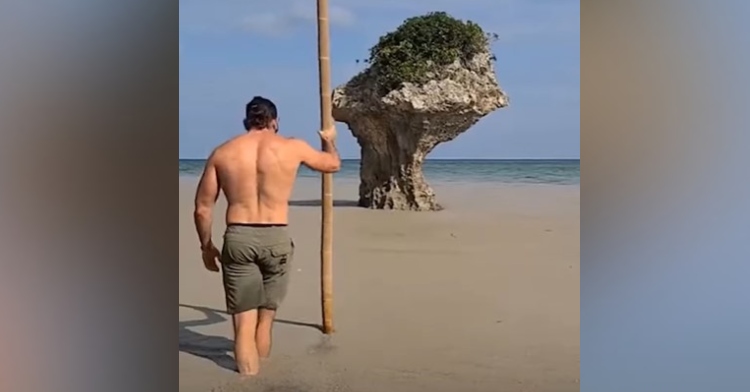 a man who goes by kylesthenix on tiktok using a large stick as he walks through pumice stone filled water on a beach in okinawa, japan after a volcano eruption