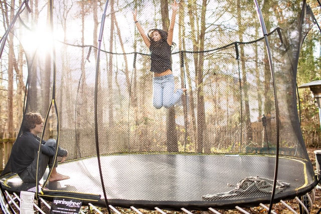 a woman jumping high on a trampoline as a man sits nearby on the trampoline and watches