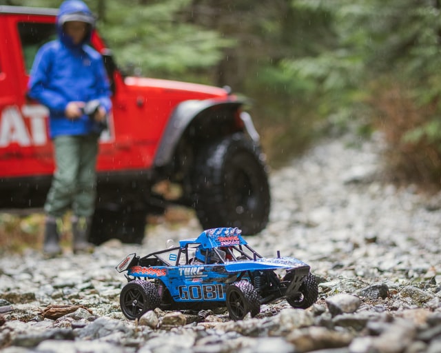 closeup of a blue remote control car as someone stands by a vehicle in the distance as they control it