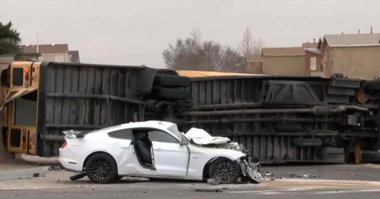 a damaged white ford mustang parked in front of a school bus that it hit that is now laying on its side