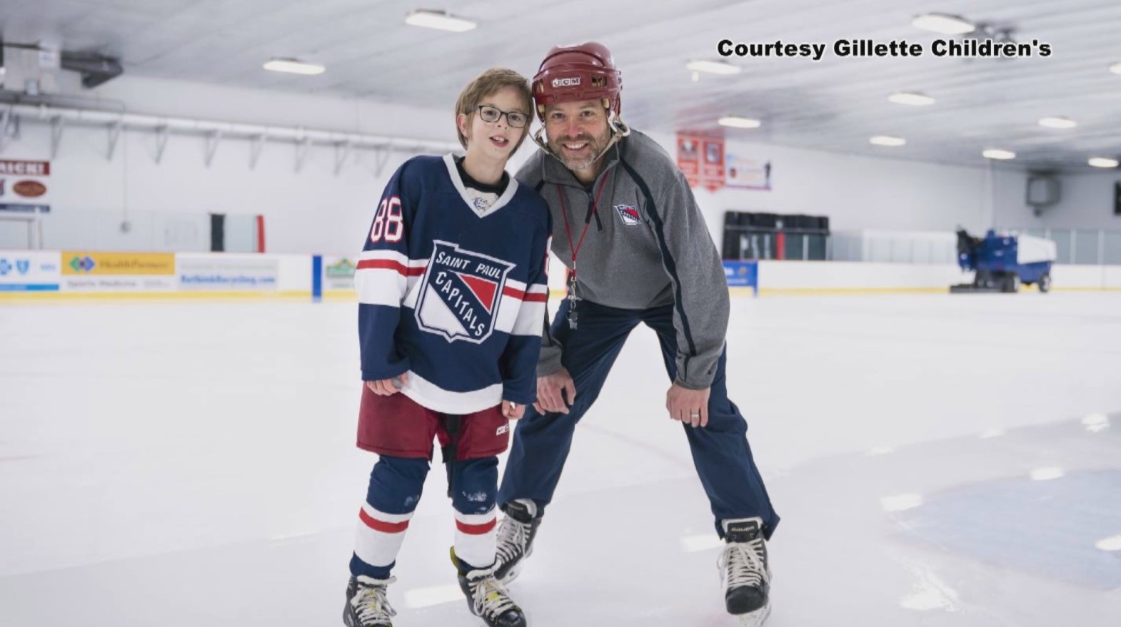 13-year-old owen nei wearing his saint paul capitals uniform as he smiles and poses with his dad and coach, chris nei, as they stand on the ice