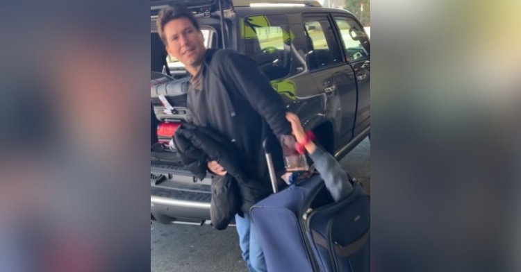 a man looking back and smiling as he pulls luggage toward an open vehicle trunk, completely unaware of his sister, kathleen, whose arm is sticking out of the luggage and is reaching for his arm
