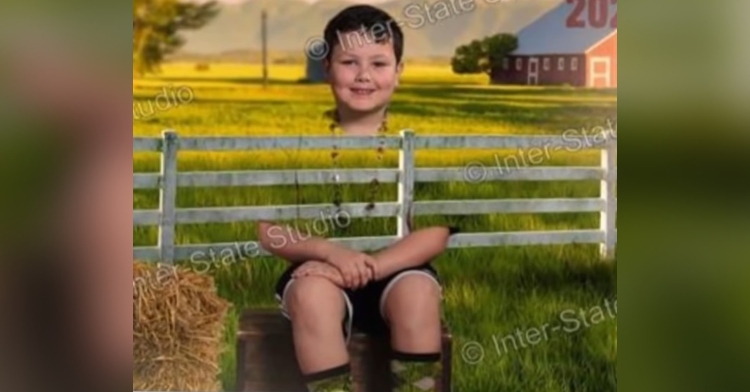 a little boy smiling as he sits and poses for a school photo at indiana elementary school with inter-state studio. because his shirt is partly green and the backdrop is a green screen, he looks partly transparent, making the fence background visible