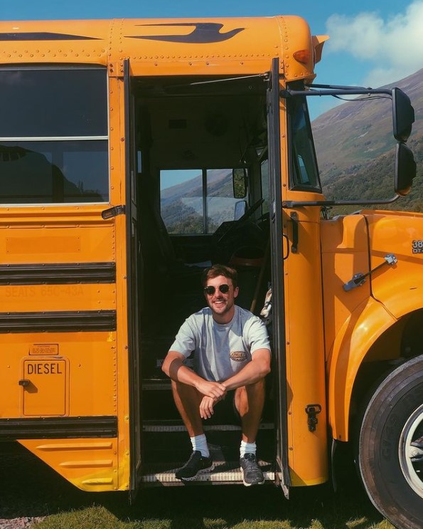 a man named angus luff smiling as he sits on the steps of the american school bus he renovated 