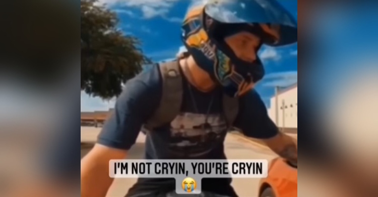screenshot of footage of a man on a motorcycle stopping to speak with a woman driving a car who almost hit him that is captioned with “I’m not cryin, you’re cryin”